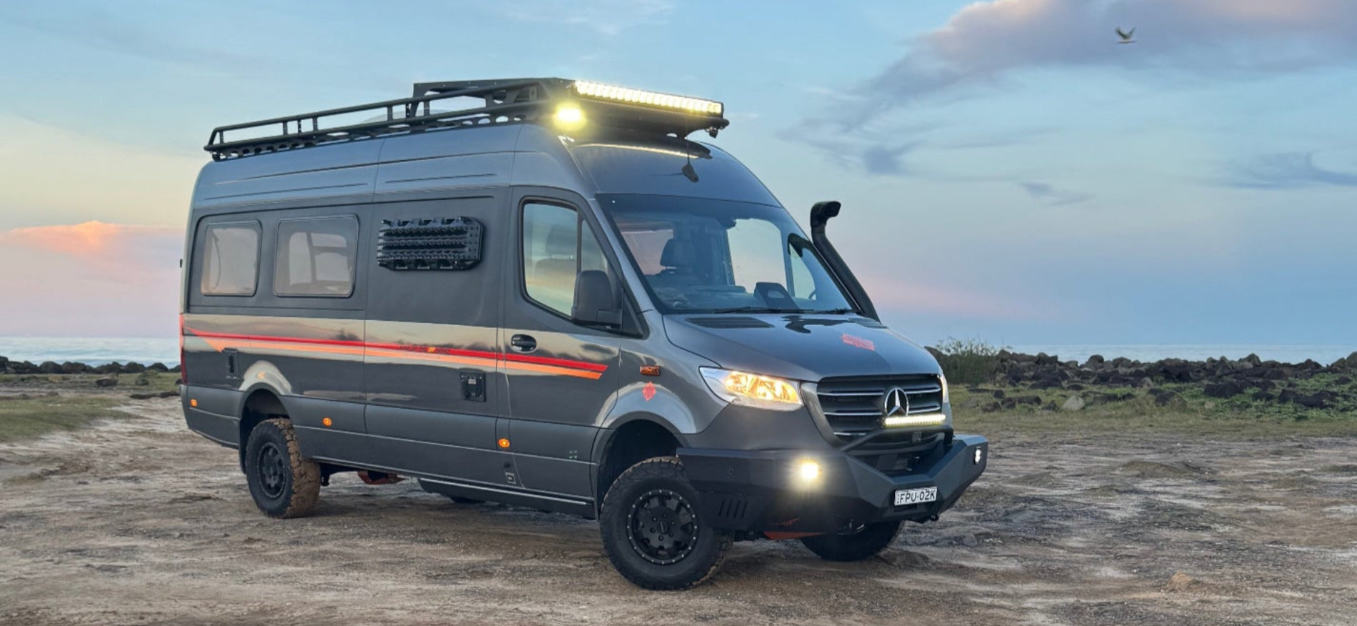 Gray camper van parked near a puddle with a sunset sky